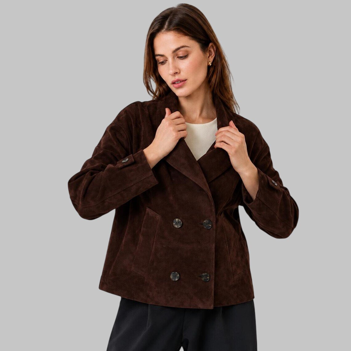 A woman wearing a dark brown suede leather double-breasted jacket over a cream top and black pants, posing against a solid gray studio background.