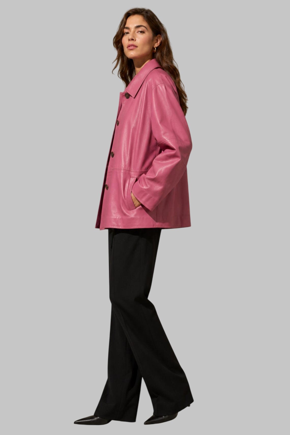 A woman modeling a shiny pink leather jacket with black wide-leg trousers, standing against a solid gray background.