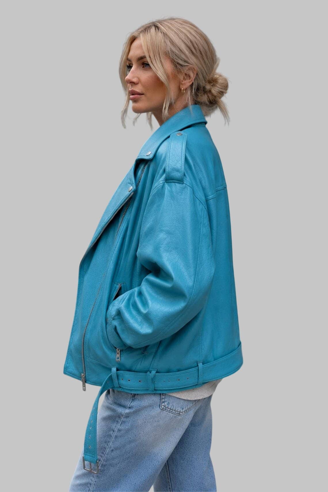 A woman wearing a bright turquoise blue leather moto jacket and light wash jeans, shown in profile against a solid gray studio background.