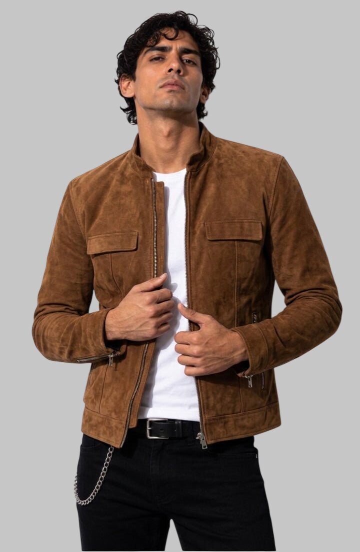 A man wearing a brown suede jacket over a white t-shirt and black pants, standing against a solid gray background.