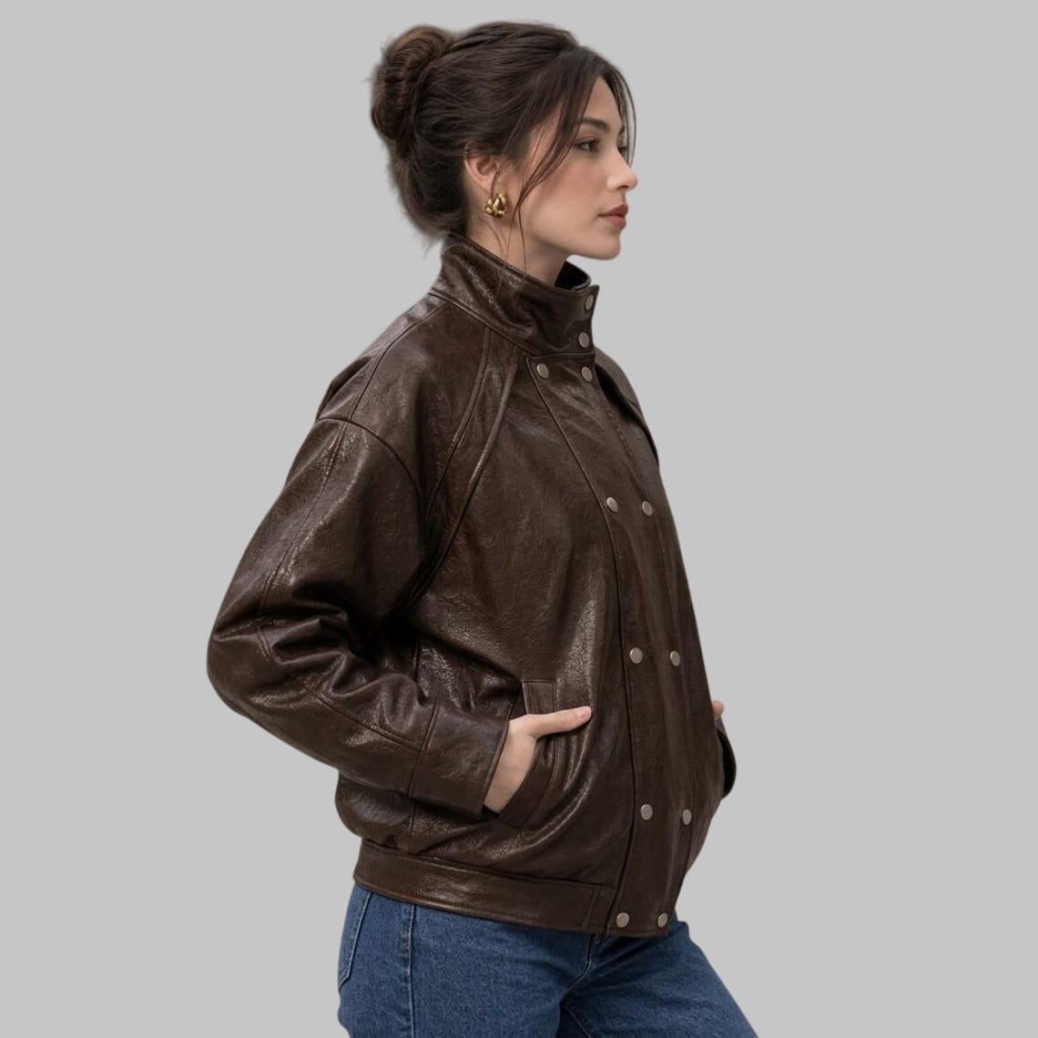 A woman wearing a dark brown leather jacket with a high collar and silver snap buttons, paired with blue jeans, shown from a side profile against a solid gray background.