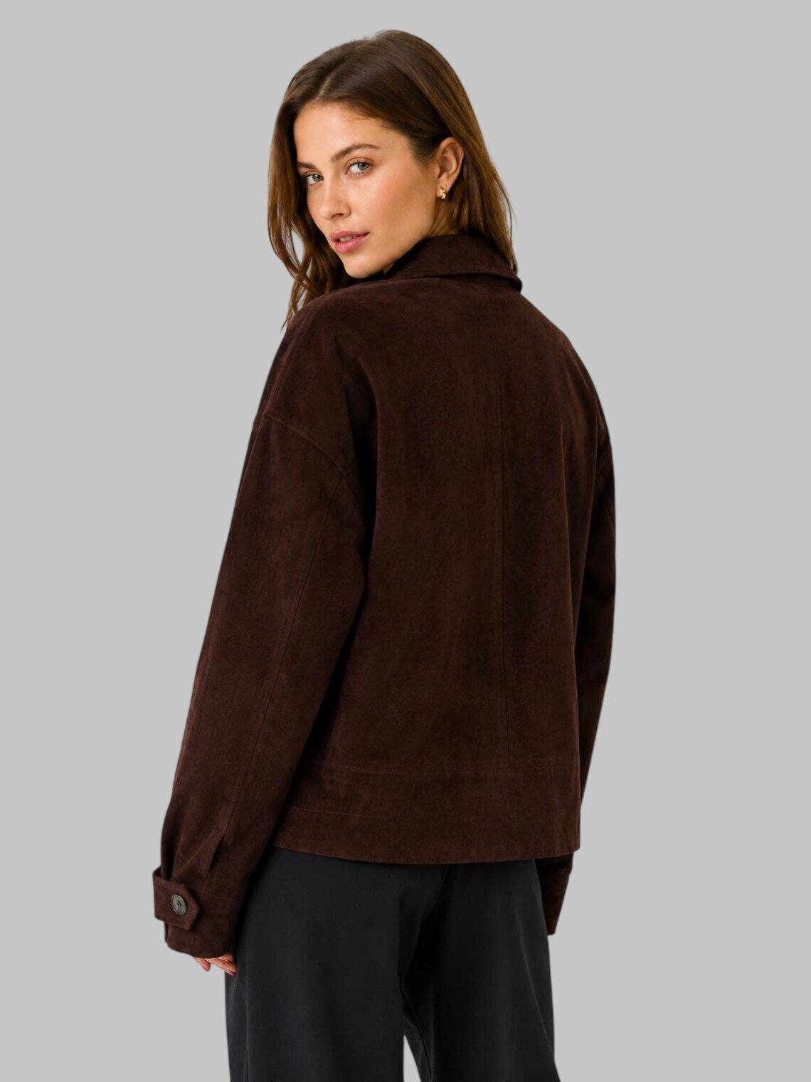 A woman wearing a dark brown suede leather jacket, shown from a three-quarters back view against a solid gray studio background.
