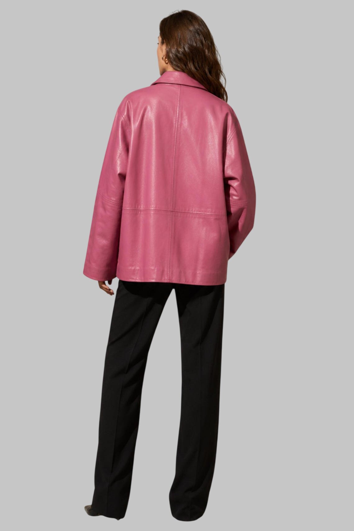 A woman shown from the back wearing a pink  leather jacket and black trousers, photographed against a solid gray studio background.