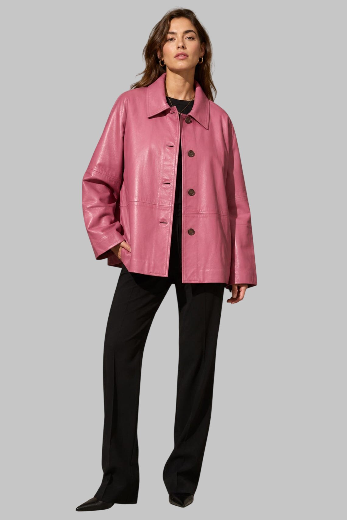 A woman modeling a pink leather jacket with a collar and front buttons, paired with black pants, against a solid gray background.