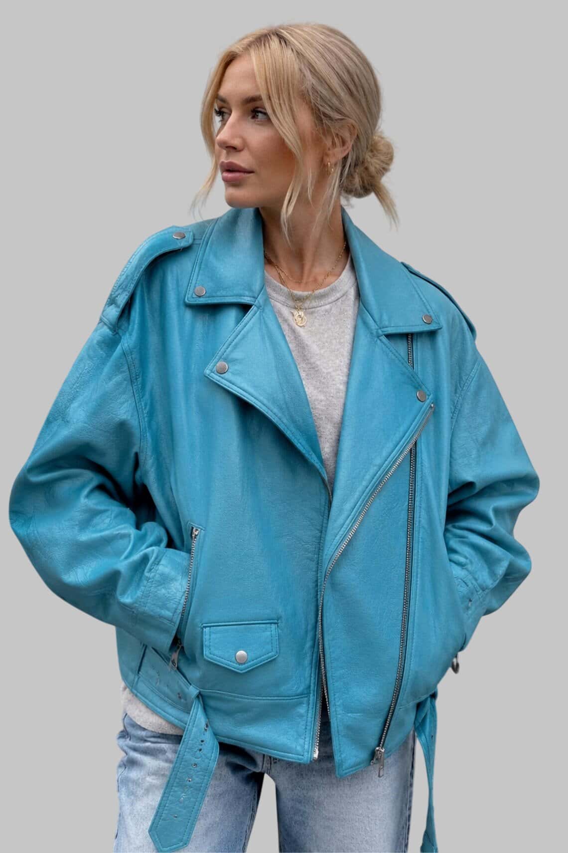A woman wearing a bright turquoise blue leather moto jacket with silver zippers and snap details, styled over a light gray t-shirt and light-wash jeans, set against a solid gray background.